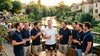 Un groupe d'amis en polos assortis rit sur une terrasse au bord de la Marne, le futur marié au centre, verres à la main